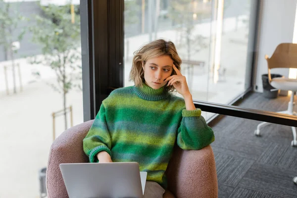 Thoughtful young woman in sweater sitting with laptop in lobby of hotel in Barcelona — стокове фото