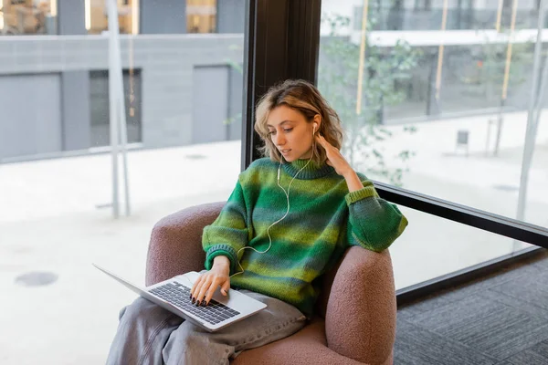 Smiling young woman in sweater and wired earphones using laptop while sitting in lobby of hotel in Barcelona — Foto stock