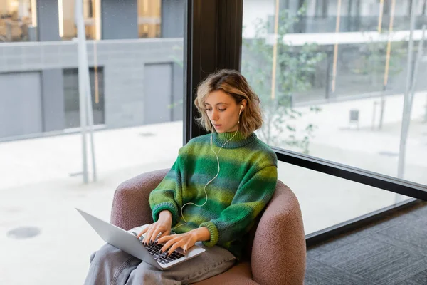 Young freelancer in sweater and wired earphones using laptop while sitting in lobby of hotel in Barcelona — Stock Photo