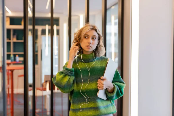Mujer rubia en suéter verde ajustando auriculares mientras sostiene el ordenador portátil en el vestíbulo del hotel en Barcelona - foto de stock