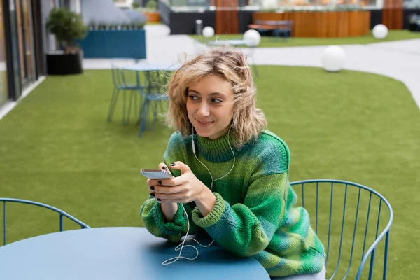 Mujer joven feliz en suéter verde escuchando música en auriculares con cable y sosteniendo el teléfono inteligente - foto de stock