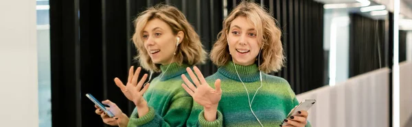 Happy woman listening music in wired earphones and holding smartphone while waving hand near mirrors in hotel, banner — Foto stock