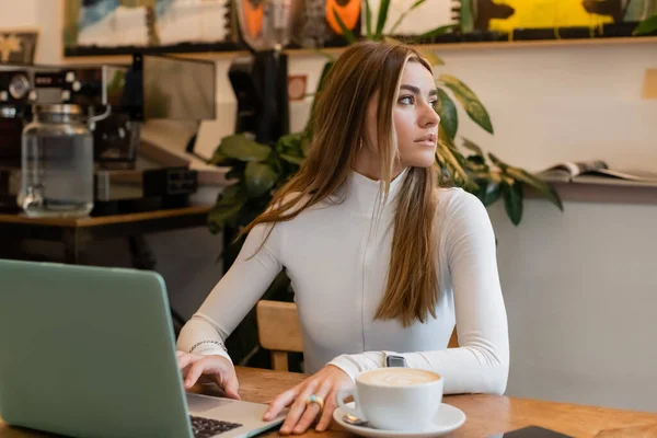 Jeune femme assise à côté de la tasse avec cappuccino et ordinateur portable sur la table dans un café à Vienne — Photo de stock