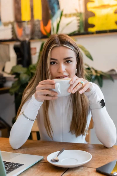 Jovem mulher bebendo cappuccino perto de laptop na mesa no café em Viena — Fotografia de Stock