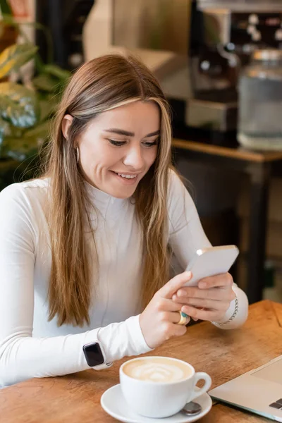 Веселый блоггер, пользующийся смартфоном рядом с ноутбуком рядом с чашкой латте в венском кафе — стоковое фото