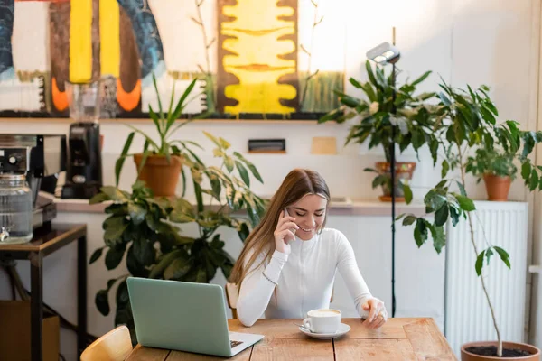 Blogger allegro che parla su smartphone vicino a computer portatile vicino a tazza di cappuccino in caffè — Foto stock