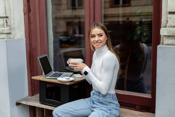 Alegre freelancer sosteniendo taza de café cerca de la computadora portátil con pantalla en blanco en la cafetería mientras está sentado al aire libre en Viena - foto de stock