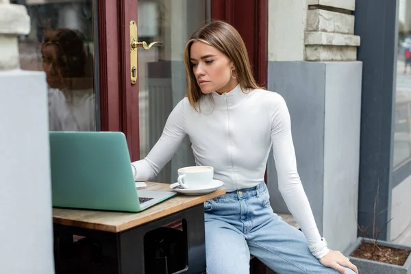 Молода жінка, яка використовує ноутбук біля чашки кави, сидячи у кафе на відкритому повітрі у Відні. — стокове фото