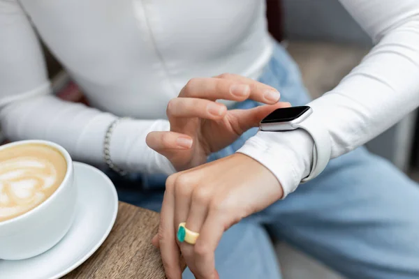 Cropped view of woman touching display on smart watch near cup of cappuccino — Stock Photo