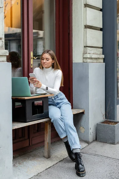 Mujer alegre usando teléfono inteligente cerca de la computadora portátil y una taza de café mientras está sentado en la cafetería en Viena - foto de stock
