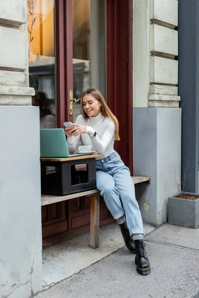 Cheerful woman using smartphone near laptop and cup of coffee while sitting in cafe on street in Vienna — Stock Photo