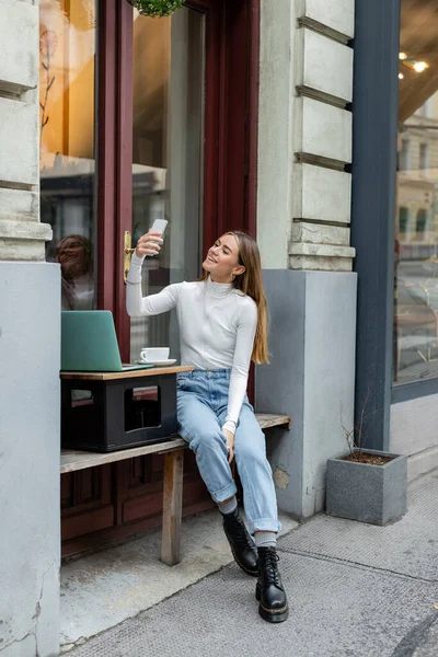 Mujer alegre tomando selfie cerca del ordenador portátil y la taza de café mientras está sentado en la cafetería en la calle en Viena - foto de stock