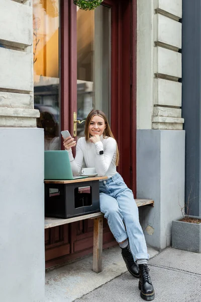 Щаслива молода жінка, яка тримає смартфон біля ноутбука і чашку кави, сидячи у кафе на вулиці у Відні. — стокове фото