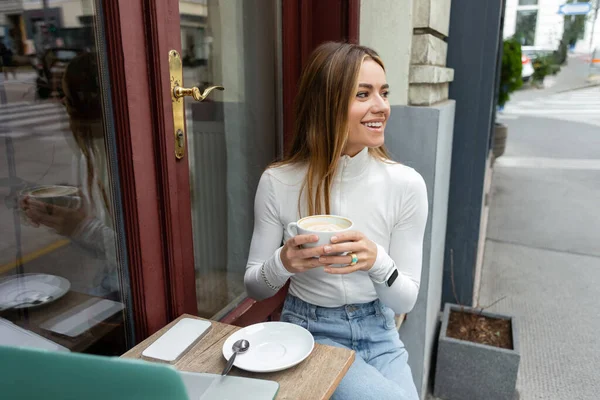 Donna spensierata che tiene in mano una tazza di cappuccino mentre siede vicino a gadget sul tavolo in un caffè in strada a Vienna — Foto stock