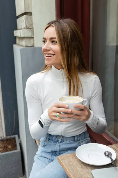 Mujer despreocupada sosteniendo taza de café con leche mientras está sentado en la cafetería al aire libre en la calle en Viena - foto de stock
