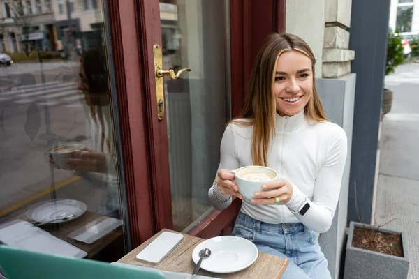 Happy woman holding cup of cappuccino while sitting at bistro table with gadgets in cafe on street in Vienna — Stock Photo