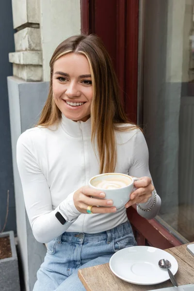 Happy woman holding cup of cappuccino while sitting at bistro table on street in Vienna — Stock Photo