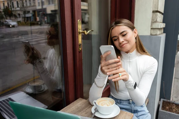 Zufriedene Frau hält Smartphone neben Tasse Latte auf Tisch, während sie in Wiener Café sitzt — Stockfoto