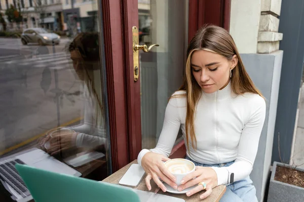 Молодая женщина смотрит на чашку с капучино, сидя за столом бистро возле гаджетов в кафе в Вене — стоковое фото