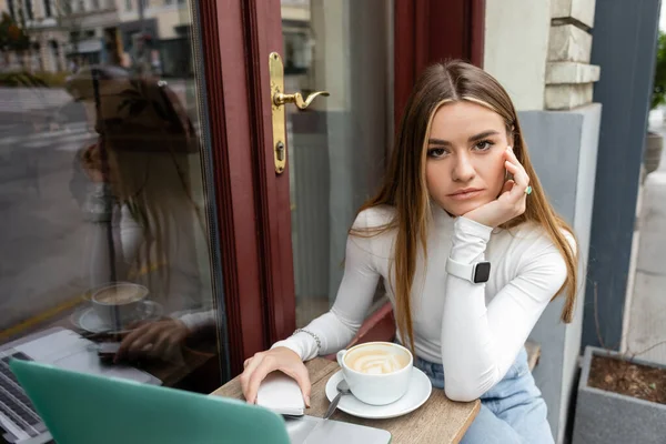 Jovem mulher segurando smartphone perto da xícara com cappuccino enquanto sentado na mesa de bistrô perto do laptop no café em Viena — Fotografia de Stock