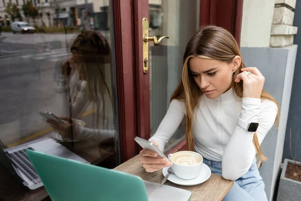 Freelancer enfocado usando smartphone cerca de la taza con cappuccino mientras está sentado en la mesa del bistro cerca del portátil en Viena - foto de stock