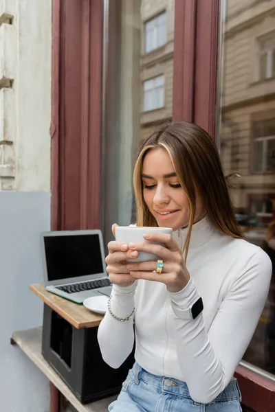 Smiling woman holding cup of coffee while sitting in outdoor cafe next to laptop with blank screen in Vienna — Stock Photo