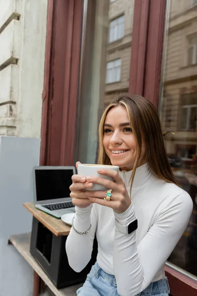 Happy woman holding cup of coffee while sitting in outdoor cafe next to laptop with blank screen in Vienna — Stock Photo