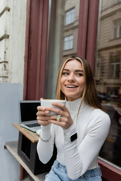Pleased woman holding cup of coffee while sitting in outdoor cafe next to laptop with blank screen in Vienna — Stock Photo