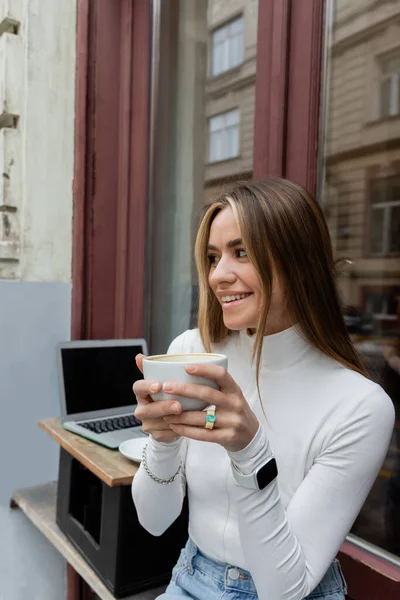 Pretty woman holding cup of coffee while sitting in outdoor cafe next to laptop with blank screen in Vienna — Stock Photo