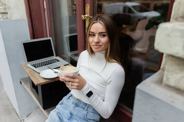 Young woman holding cup of cappuccino while sitting in outdoor cafe next to laptop with blank screen in Vienna — Stock Photo