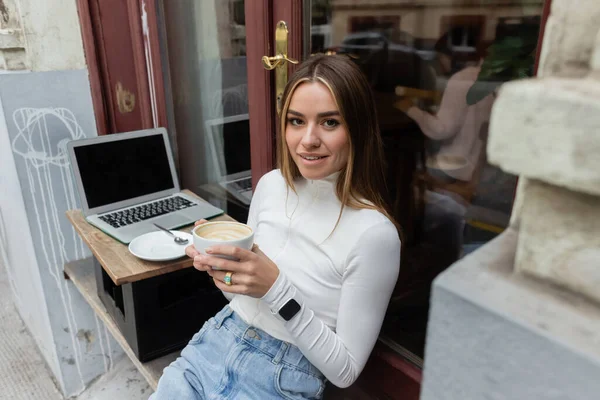 Encantadora mulher segurando xícara de cappuccino enquanto sentado no café ao ar livre ao lado do laptop com tela em branco em Viena — Fotografia de Stock