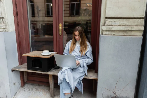 Young freelancer in blue trench coat using laptop while sitting next to cup of coffee in outdoor cafe in Vienna — Stock Photo