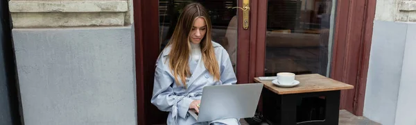 Young freelancer in blue trench coat using laptop while sitting next to cup of coffee in outdoor cafe in Vienna, banner — Stock Photo