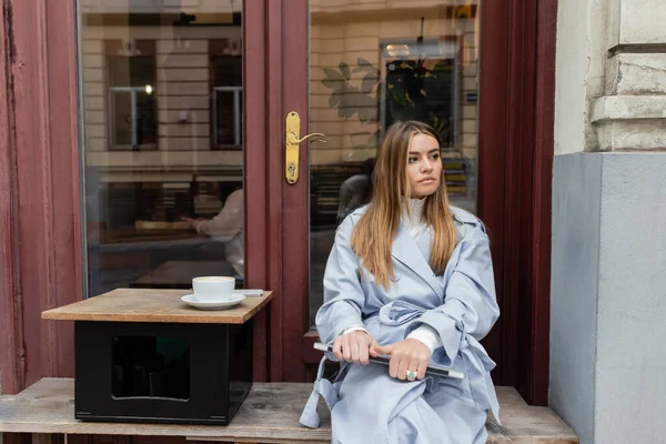 Молода жінка в синьому плащі, тримаючи ноутбук, сидячи біля чашки кави у відкритому кафе у Відні. — стокове фото