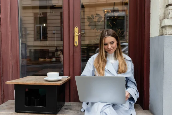 Cheerful freelancer in blue trench coat using laptop while sitting next to cup of coffee in outdoor cafe in Vienna — Stock Photo