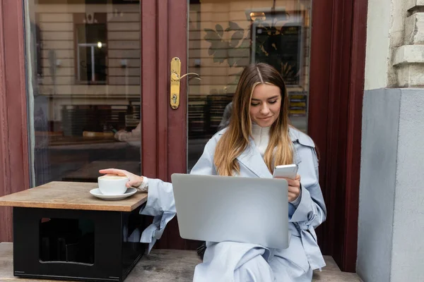 Jovem mulher em casaco de trincheira azul usando smartphone perto de laptop enquanto sentado ao lado da xícara de café no café ao ar livre em Viena — Fotografia de Stock