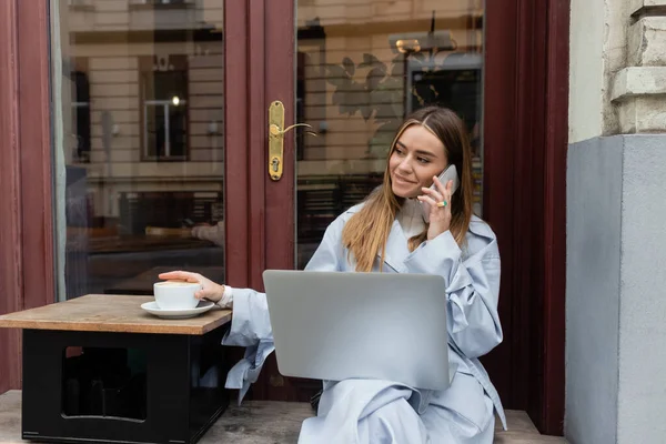 Femme gaie en trench coat bleu parlant sur smartphone et assis à côté d'une tasse de café dans un café en plein air à Vienne — Photo de stock