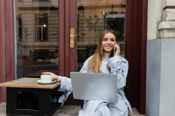 Femme heureuse en trench coat bleu parlant sur smartphone et assis près d'une tasse de café dans un café extérieur à Vienne — Photo de stock