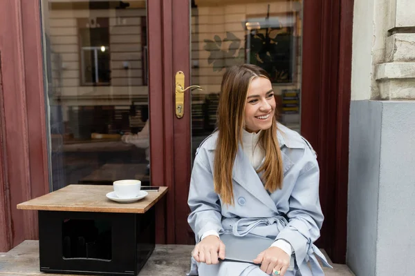 Heureuse jeune femme en trench coat bleu tenant ordinateur portable tout en étant assis à côté d'une tasse de café dans un café extérieur à Vienne — Photo de stock