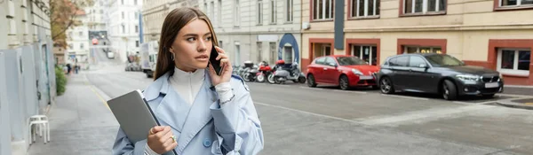Jeune femme en trench coat bleu parlant sur smartphone tout en tenant un ordinateur portable dans la rue à Vienne, bannière — Photo de stock