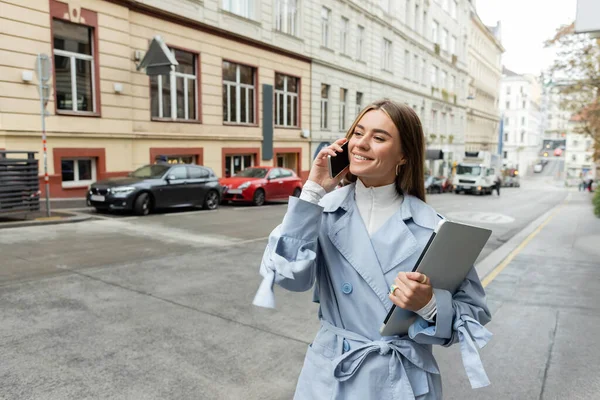 Mujer alegre en gabardina azul hablando en el teléfono inteligente mientras sostiene el ordenador portátil en la calle en Viena - foto de stock