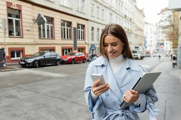 Joyful woman in blue trench coat using smartphone while holding laptop on street in Vienna — Stock Photo