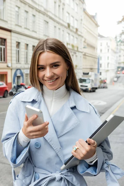 Smiling woman in blue trench coat using smartphone while holding laptop on street in Vienna — Stock Photo