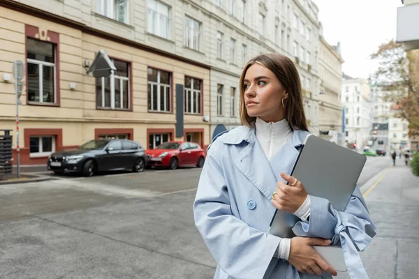 Pretty woman in blue trench coat holding smartphone and laptop while walking on street in Vienna — Stock Photo