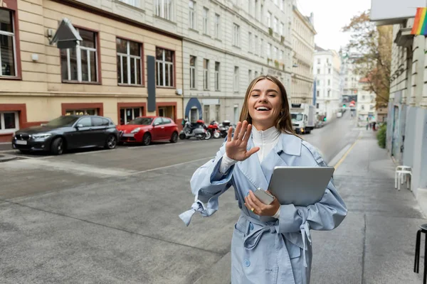 Happy woman in blue trench coat holding smartphone and laptop while waving hand on street in Vienna — Stock Photo