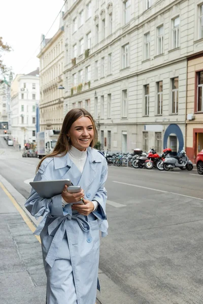 Mujer feliz en gabardina de moda sosteniendo teléfono inteligente y portátil mientras camina por la calle en Austria - foto de stock