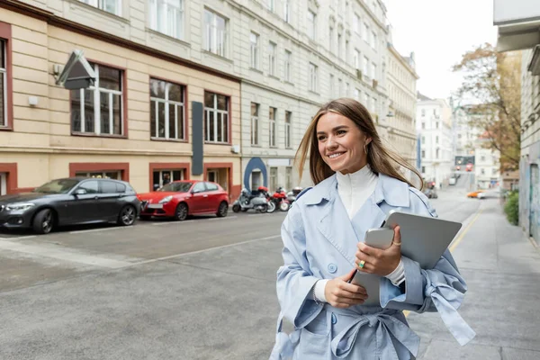 Pleased woman in stylish trench coat holding smartphone and laptop while walking on street in Vienna — Stock Photo