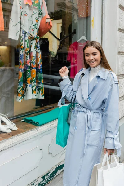 Mujer despreocupada en elegante gabardina de pie con bolso y bolsas de compras cerca del escaparate de la tienda boutique en Viena - foto de stock
