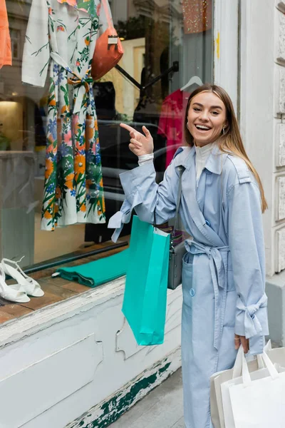 Mujer despreocupada en elegante gabardina de pie con bolsas de compras y apuntando a la vitrina de la tienda boutique en Viena - foto de stock
