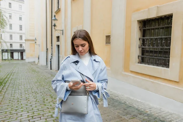 Young woman in blue trench coat and handbag using smartphone on street in Vienna — Stock Photo